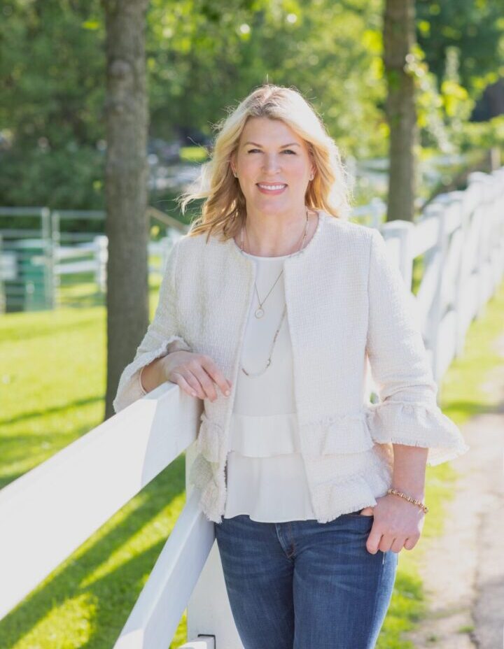 A woman in white jacket standing on grass near trees.